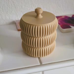 Könnte beinhalten: Ein hellbeiger, zylindrischer Aufbewahrungsbehälter mit stapelbarem Design, bestehend aus drei gerippten, abgerundeten Abschnitten. Der Behälter verfügt über einen flachen, runden Deckel mit einem kleinen, zentralen Knauf zum einfachen Öffnen.