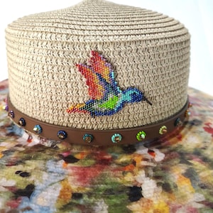 May include: A straw hat with a flat crown and a wide brim featuring a multicolored floral pattern. A brown band encircles the base, adorned with small, colorful, circular beaded accents and a central beaded hummingbird in vibrant rainbow hues.