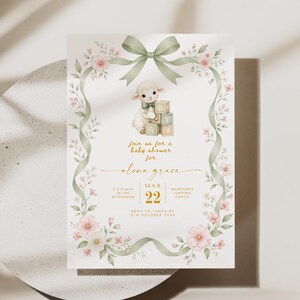 May include: A rectangular baby shower invitation card featuring a watercolor illustration of a lamb with alphabet blocks, framed by a sage green ribbon and pink floral vines. The text includes details for Alena Grace on March 22.
