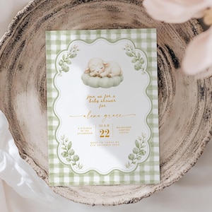 May include: A rectangular baby shower invitation card featuring a green and white gingham border, a central illustration of a sleeping lamb on a cushion, and gold text reading Join us for a baby shower for Alena Grace, March 22, 8 o'clock in the afternoon.