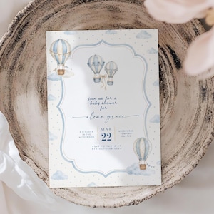 Puede incluir: Una tarjeta de invitaci&oacute;n rectangular para baby shower con ilustraciones en acuarela de globos aerost&aacute;ticos de color azul claro y blanco y nubes sobre un fondo blanco con lunares. El texto dice: Join us for a baby shower for Alena Grace, Mar 22.