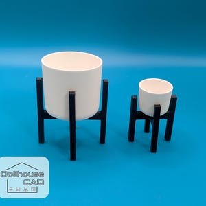 Op de afbeelding: Twee witte cilindervormige plantenbakken op zwarte metalen standaards. De grotere plantenbak is hoger dan de kleinere. De standaards hebben een minimalistisch ontwerp. De achtergrond is effen blauw. Het logo "Dollhouse CAD" staat linksonder.