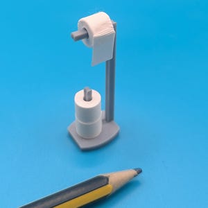 May include: A miniature gray toilet paper holder with a roll of white toilet paper and two additional rolls. A blue and yellow pencil is in the foreground against a blue background.