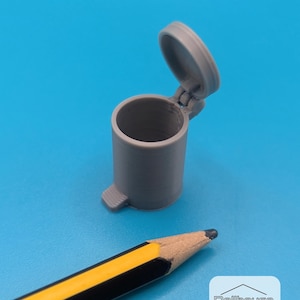 May include: A small, gray miniature trash can with a hinged lid, open. The trash can has a foot pedal detail. A yellow and black pencil is in the foreground. The background is a solid blue. The image includes the text "Dollhouse CAD".