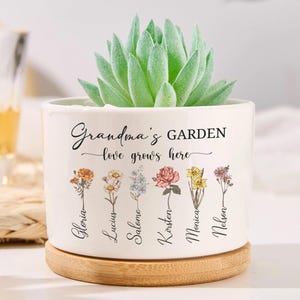May include: A white ceramic plant pot with a succulent. The pot has the words "Grandma's GARDEN love grows here" and floral illustrations. The names "Gloria, Lucius, Salome, Kirsten, Monica, Nelson" are below the flowers. The pot sits on a wooden base.
