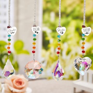 Könnte beinhalten: Vier personalisierte Kristall-Sonnenfänger mit herzförmigen Anhängern, die jeweils den Namen eines Haustiers tragen: Charlie, Oscar, Shadow und Shelby. Die Anhänger sind mit einer Kette aus bunten Perlen und einem Kristallprisma verbunden. Der Hintergrund zeigt eine verschwommene Gartenszene.