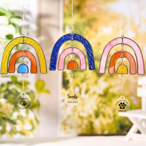 May include: Three colorful stained glass rainbow suncatchers. Each rainbow has a different color scheme: yellow, orange, and blue; blue, pink, and orange; and pink, orange, and yellow. Each has a charm with a paw print and text.