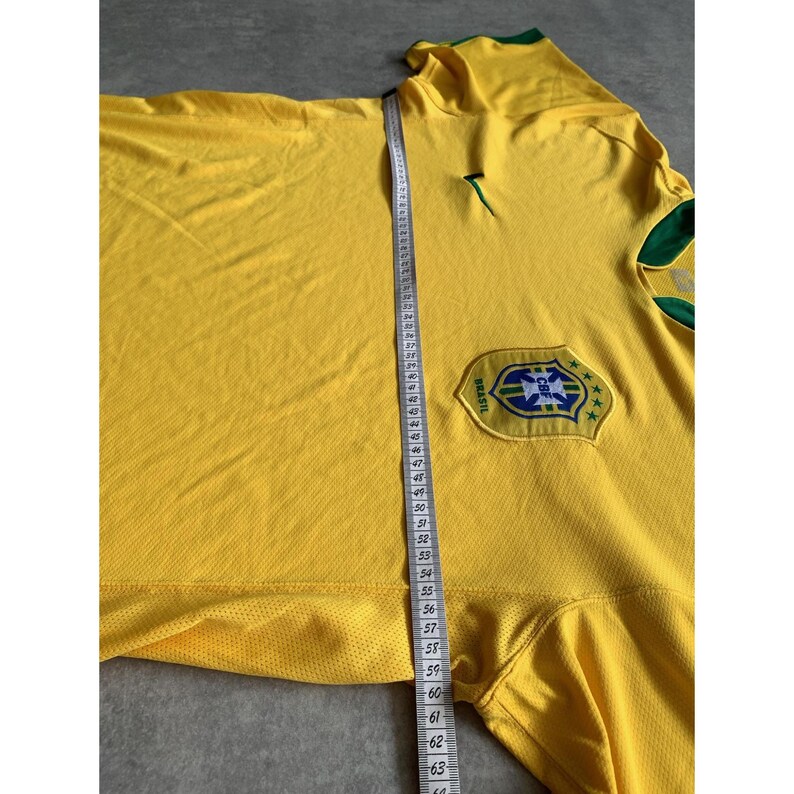 May include: A yellow Brazil soccer jersey with green and blue accents. The jersey features the CBF logo and a green collar. A measuring tape shows the length of the jersey. The jersey is made of a breathable fabric.