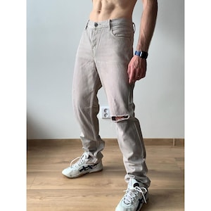 May include: Light beige denim jeans with a distressed knee detail. The jeans have a classic five-pocket design and a button and zipper closure. The model is wearing white and black sneakers.