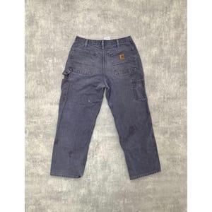 May include: Faded blue work pants with multiple pockets and a brown logo patch. The pants have a relaxed fit and are made of durable material. The back features two large pockets and a hammer loop.