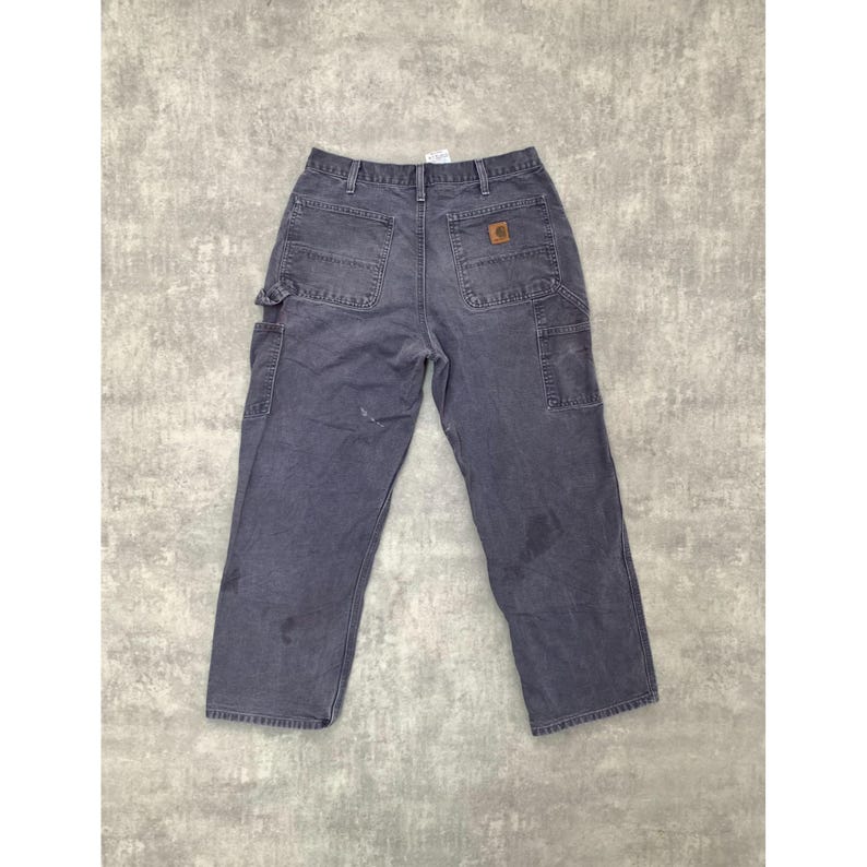 May include: Faded blue work pants with multiple pockets and a brown logo patch. The pants have a relaxed fit and are made of durable material. The back features two large pockets and a hammer loop.