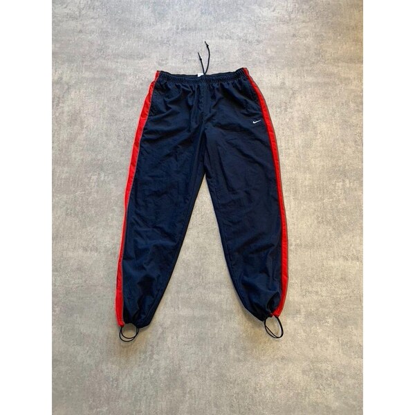 May include: Navy blue tracksuit bottoms with red side stripes. The trousers have an elasticated waist with a drawstring and a small white Nike logo. The bottoms have drawstrings. The trousers are made of a lightweight material.