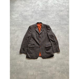 May include: A brown and blue plaid blazer with an orange interior. The blazer has two front pockets, brown buttons, and a brown elbow patch. The jacket is laid flat on a gray surface.