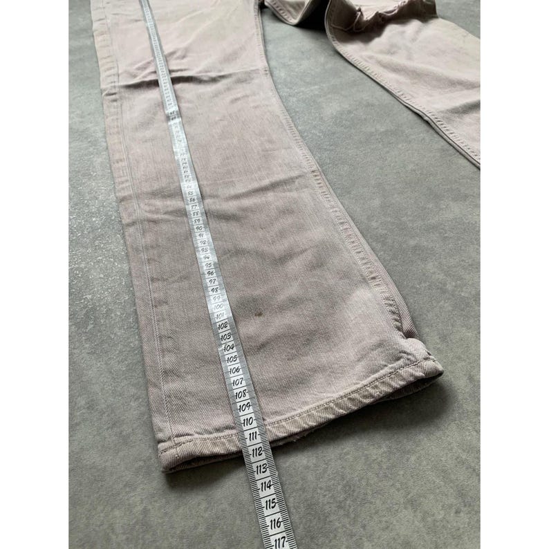 May include: Light brown denim pants with a straight leg design. A measuring tape is laid along the length of the pants. The pants are made of a sturdy material and appear to be in good condition.