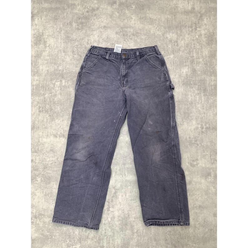 May include: Faded blue work pants with multiple pockets and a button closure. The pants show signs of wear and tear, with some white paint marks. The pants are made of a durable material, suitable for work or casual wear.
