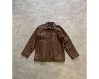 Può includere: Giacca in pelle marrone con collo e chiusura a zip. La giacca ha una tasca sul petto e maniche lunghe. La giacca è esposta su una superficie grigia.