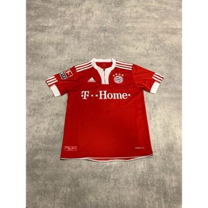 May include: Red soccer jersey with white accents, featuring the Adidas logo and the text "T. Home." The jersey has a white collar and sleeve cuffs with three white stripes. A team emblem is on the chest, and a Bundesliga logo is on the sleeve.