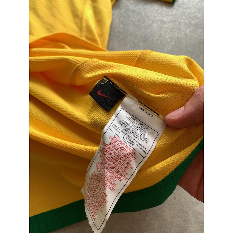 May include: A close-up of a yellow and green athletic jersey. The jersey has a Nike logo and a tag with text. The jersey is made of a breathable, textured fabric. The collar and sleeve cuffs are green.