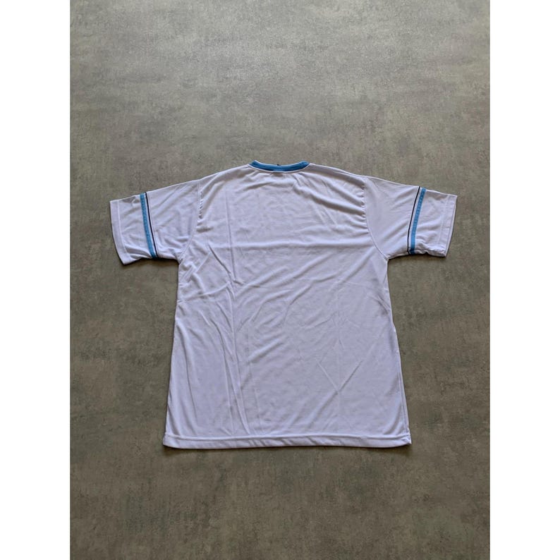 May include: White t-shirt with light blue trim on the collar and sleeves. The shirt is made of a lightweight material and has short sleeves. The shirt is laid flat on a gray surface.