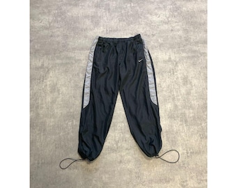 Puede incluir: Pantalones de chándal Nike negros con rayas laterales grises y un pequeño logotipo de Nike. Los pantalones tienen cordones en los tobillos y la cintura. Los pantalones están hechos de un material ligero.