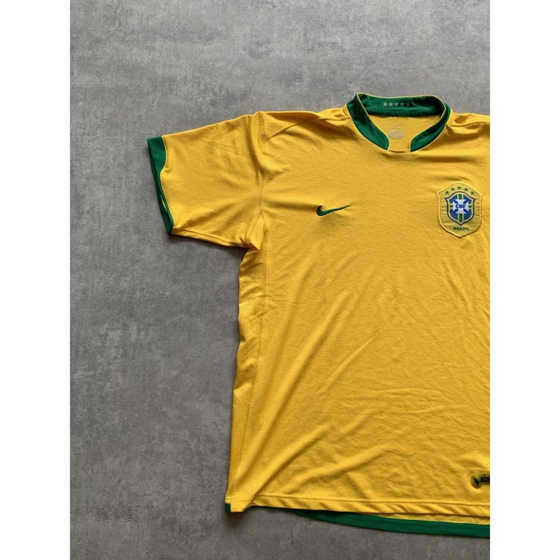 May include: A yellow Brazil soccer jersey with green trim on the collar and sleeves. The jersey features the Nike swoosh logo in green, along with the Brazilian national team crest on the chest. The jersey is made of a lightweight material.