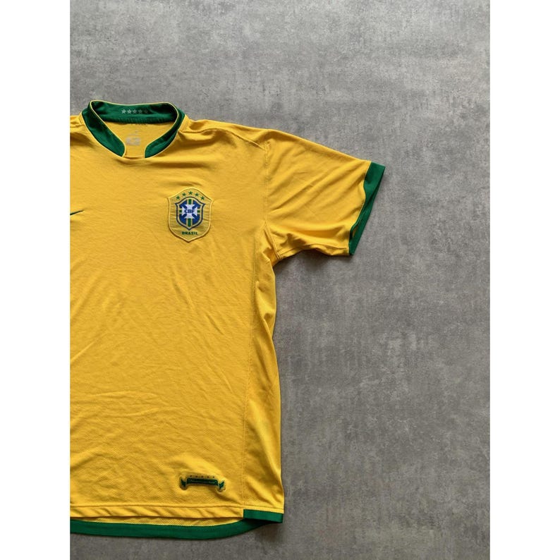 May include: A yellow soccer jersey with green trim on the collar and sleeves. The jersey features a crest with a blue and white design and the word "Brasil". The Nike logo is visible on the chest.