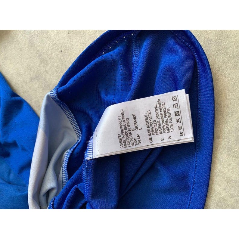 Pu&ograve; includere: Primo piano di una maglia sportiva blu con un'etichetta bianca. L'etichetta include testo in pi&ugrave; lingue, tra cui "Made in Philippines" e "100% Poliestere". La maglia &egrave; realizzata in tessuto traspirante.