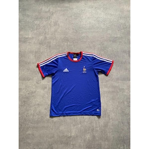 May include: Blue Adidas football shirt with white shoulder stripes and red trim. Includes the Adidas logo and a small rooster emblem. Short-sleeved, crew neck design.