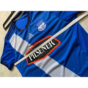 Pu&ograve; includere: Maglia da calcio blu con strisce bianche e grigie, con il logo "EMELEC" e il testo "PILSENER". Il logo Adidas &egrave; visibile. Un metro a nastro &egrave; appoggiato sulla maglia.