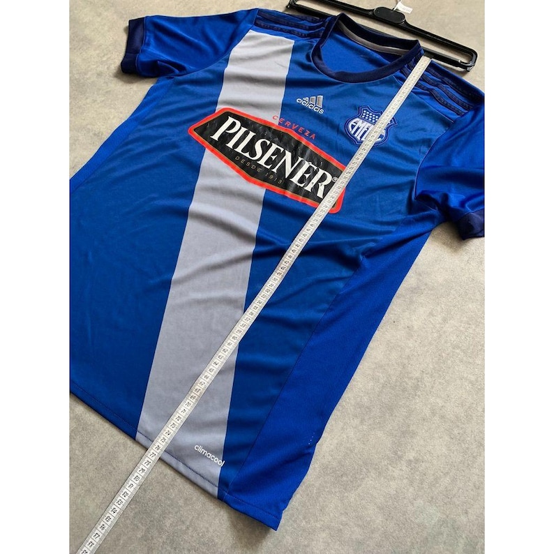Pu&ograve; includere: Maglia da calcio blu e grigia con il logo "Pilsener" e il testo "Desde 1918". La maglia ha una striscia bianca sul davanti e viene misurata con un metro a nastro. Il logo Adidas &egrave; visibile.