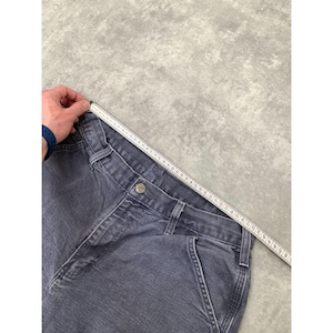 May include: A pair of blue denim work pants with a button closure and multiple pockets. A measuring tape is laid across the waistband. The pants are a faded blue color.