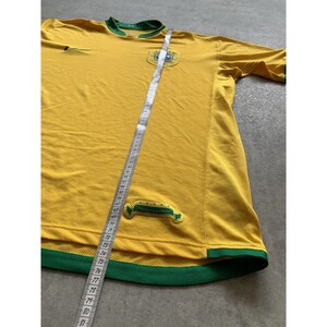 May include: Yellow soccer jersey with green trim and accents. The jersey has a crest on the chest and a Nike logo. A measuring tape is laid across the jersey. The jersey is made of a breathable material.