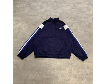May include: Navy blue Puma track jacket with white and blue stripes down the sleeves. The jacket has a full zipper and a small Puma logo on the chest. The jacket is laid flat on a gray surface.