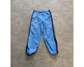 Könnte beinhalten: Hellblaue Sporthose mit dunkelblauen Seitenstreifen und Kordelzugbündchen. Die Hose hat einen elastischen Bund und ein kleines Logo am oberen linken Bein. Der Stoff scheint leicht zu sein, möglicherweise Nylon oder Polyester.
