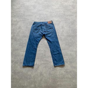 May include: Blue denim jeans with a classic five-pocket design. The jeans have a straight leg cut and a brown leather patch on the back waistband. The jeans are laid flat on a grey surface.