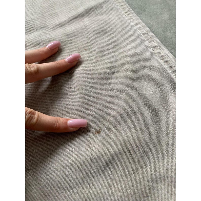 May include: Close-up of light gray denim fabric with visible texture and stitching. A hand with pink nail polish is touching the fabric, with a small brown stain visible on the denim.