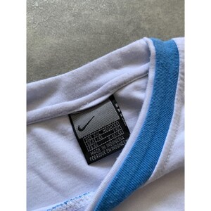 May include: Close-up of a white t-shirt with a light blue trim around the collar. A black tag with the Nike logo and text is visible. The tag includes size information and the text "MADE IN INDONESIA".