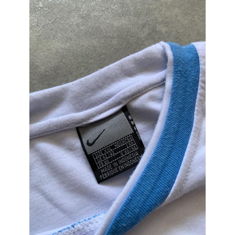 May include: Close-up of a white t-shirt with a light blue trim around the collar. A black tag with the Nike logo and text is visible. The tag includes size information and the text "MADE IN INDONESIA".