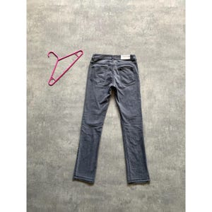 May include: A pair of gray denim jeans with a classic five-pocket design. The jeans are laid flat on a gray surface, with a pink hanger to the left. The jeans have a straight leg cut and a faded appearance.