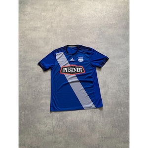 Pu&ograve; includere: Maglia da calcio blu a maniche corte con una striscia diagonale grigia e la scritta "PILSENER" in un rettangolo rosso. La maglia ha un colletto nero e il logo Adidas.