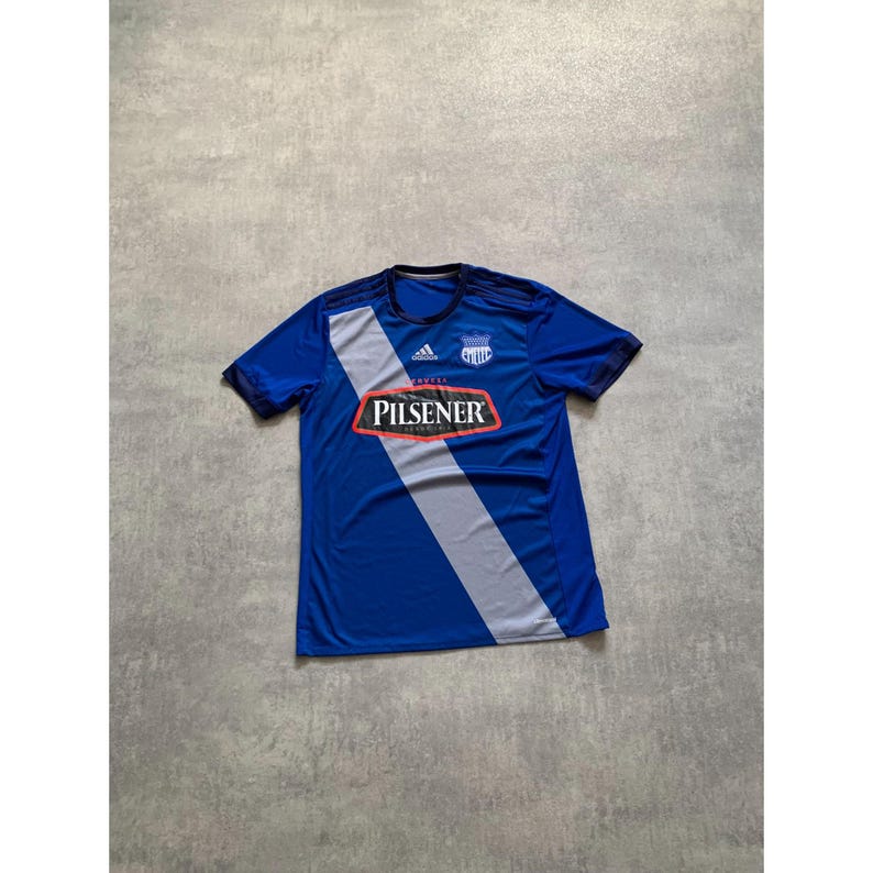 Pu&ograve; includere: Maglia da calcio blu a maniche corte con una striscia diagonale grigia e la scritta "PILSENER" in un rettangolo rosso. La maglia ha un colletto nero e il logo Adidas.