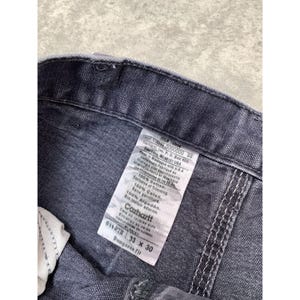 May include: Close-up of a pair of gray Carhartt Dungaree Fit work pants. The label shows the size 33 x 30, and the fabric is 100% cotton. The stitching is visible along the seams.