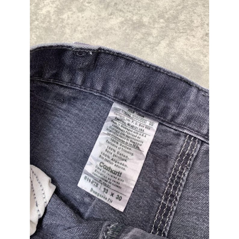 May include: Close-up of a pair of gray Carhartt Dungaree Fit work pants. The label shows the size 33 x 30, and the fabric is 100% cotton. The stitching is visible along the seams.