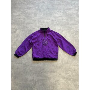 Puede incluir: Chaqueta tipo jersey morada con cuello, puños y dobladillo negros. La chaqueta presenta un patrón geométrico y un emblema circular en el pecho. Tiene cierre de cremallera de un cuarto.