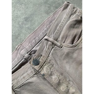 May include: Close-up of taupe-colored denim pants. The pants feature a button closure with a decorative button, a front pocket, and visible stitching details. The fabric appears to be slightly faded, giving it a vintage look.