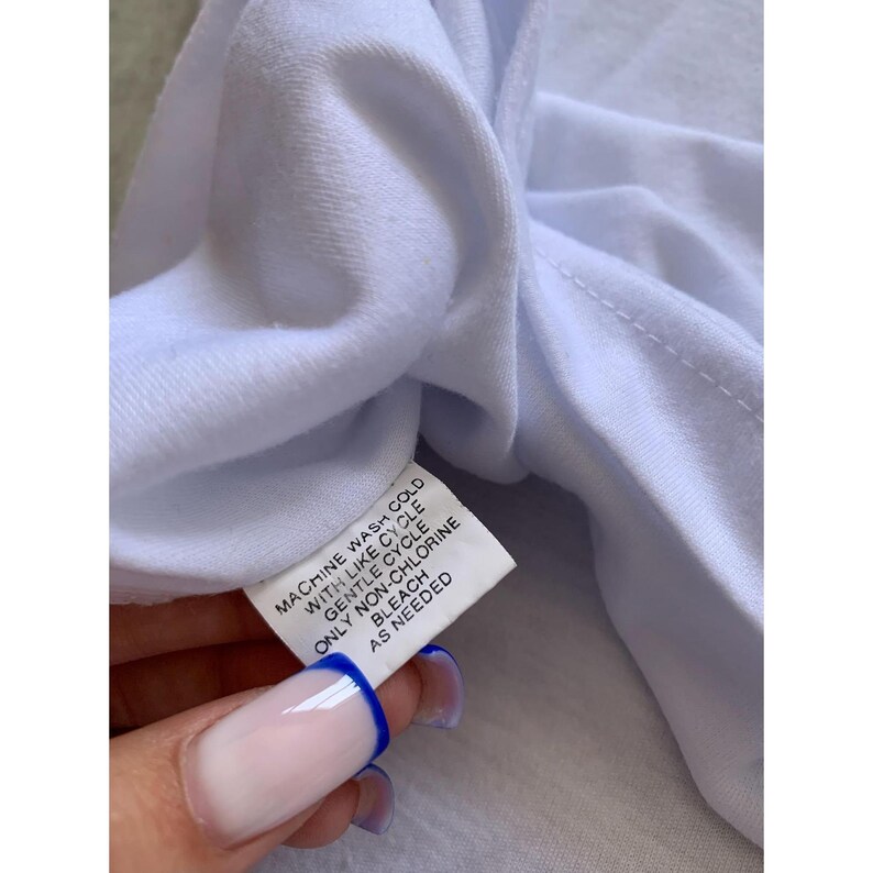 May include: Close-up of a white garment with a care label. The label includes instructions for machine washing in cold water with a gentle cycle, non-chlorine bleach, and as needed.