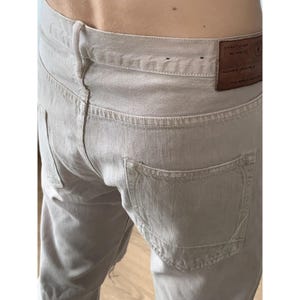 May include: Close-up of light beige denim jeans. The jeans have a classic five-pocket design with a leather patch on the back waistband. The stitching is visible throughout the garment, and the overall style is casual.