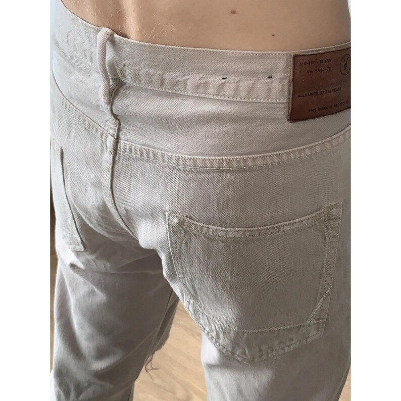 May include: Close-up of light beige denim jeans. The jeans have a classic five-pocket design with a leather patch on the back waistband. The stitching is visible throughout the garment, and the overall style is casual.