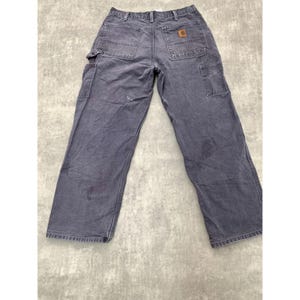 May include: Faded navy blue work pants with multiple pockets and a brown logo patch on the back. The pants have a relaxed fit and show signs of wear. The Carhartt brand is visible.