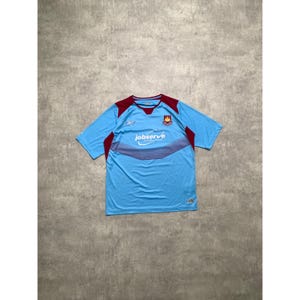 May include: A light blue short-sleeved football shirt with maroon detailing on the shoulders and neckline. The shirt has the text "jobserve" across the chest and a team crest on the upper left. The Reebok logo is visible.